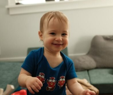 A smiling baby in a blue onesie stands indoors, used as an editorial illustration for home safety as babies begin pulling to stand.