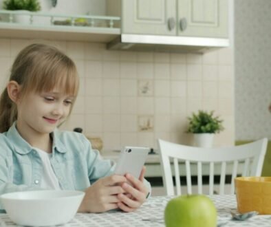 A young person uses a smartphone at a kitchen table while a parent brings breakfast.